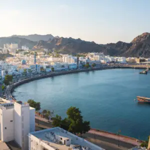 A captivating view of Muscat’s Corniche, featuring the tranquil waterfront, traditional architecture, and the historic Muttrah harbor set against rugged mountains. A dhow peacefully floats in the Gulf of Oman, reflecting the city’s rich maritime heritage.